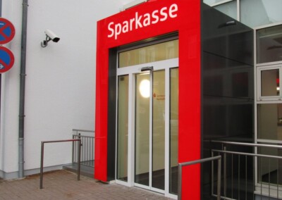 Der Eingang einer Sparkassenfiliale mit rotem Rahmen und automatischen Glastüren, neben einem weißen Gebäude.