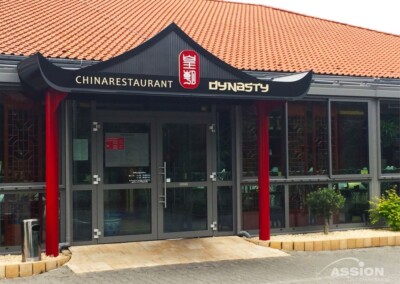 Eingang eines chinesischen Restaurants namens "Dynasty" mit traditionellen architektonischen Elementen und Glastüren unter einem roten Ziegeldach.