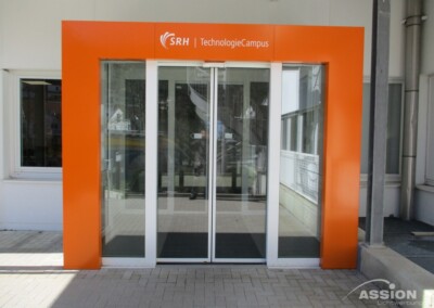 Automatische Eingangstüren aus Glas mit orangefarbenem Rahmen und dem Schriftzug "SRH TechnologieCampus" darüber, am Eingang eines modernen Gebäudes.