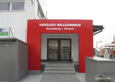 Eingang zu einem Gebäude mit einem roten Schild "HERZLICH WILLKOMMEN Ausstellung | Verkauf" über der Tür, das auf einen Ausstellungs- und Verkaufsbereich hinweist.