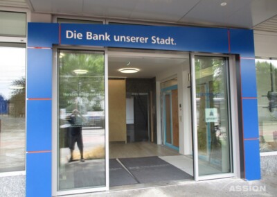 Eingang einer modernen Bankfiliale mit dem deutschen Schild "Die Bank unserer Stadt". Automatische Glastüren und eine Willkommensmatte sind zu sehen.