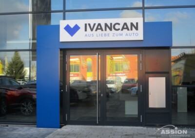 Der Eingang eines modernen Gebäudes mit einer Glasfassade zeigt ein Schild mit der Aufschrift "IVANCAN AUS LIEBE ZUM AUTO" über doppelten Glastüren.