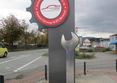 Großes Schild in Form eines Zahnrads und eines Schraubenschlüssels vor einem Geschäft, mit Text, der auf eine Autowerkstatt hinweist, in der Nähe einer Straße und eines Parkplatzes.