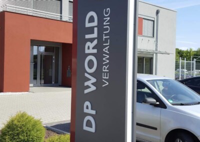 Auf einem hohen Schild vor einem Gebäude steht "DP WORLD VERWALTUNG" mit einem mehrfarbigen Logo und der Zahl 13 an der Spitze. In der Nähe sind ein weißes Auto und Sträucher zu sehen.