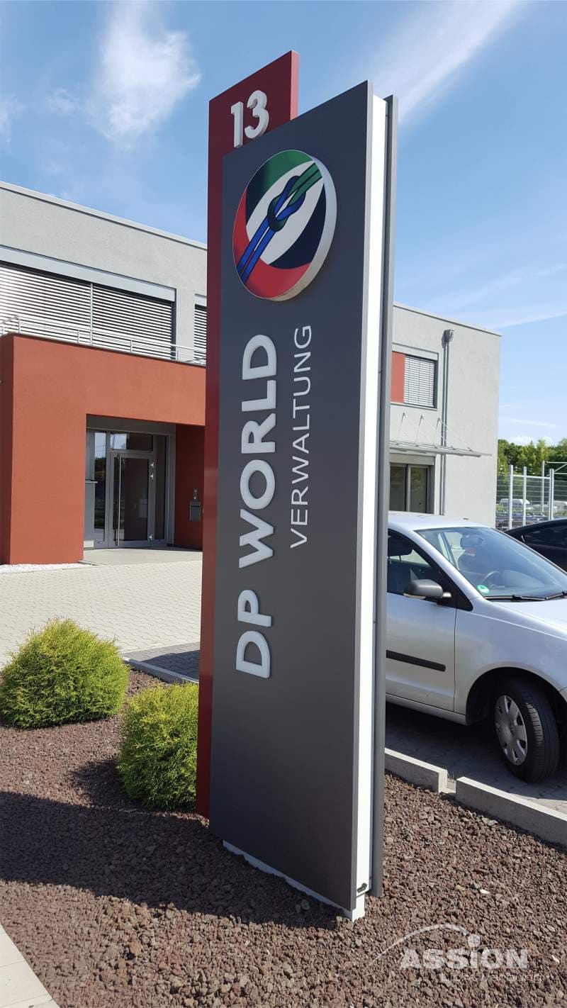 Auf einem hohen Schild vor einem Gebäude steht "DP WORLD VERWALTUNG" mit einem mehrfarbigen Logo und der Zahl 13 an der Spitze. In der Nähe sind ein weißes Auto und Sträucher zu sehen.