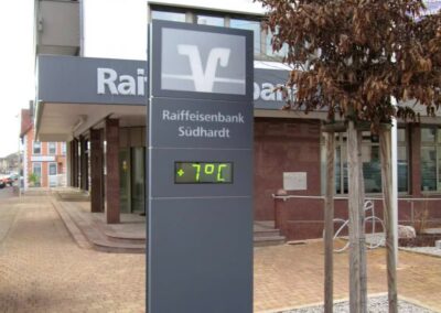 Ein digitales Schild vor der Raiffeisenbank Südhardt zeigt die Temperatur von 7°C an; im Hintergrund sind das Bankgebäude und ein Baum zu sehen.