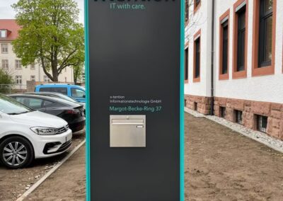 Großes rechteckiges Schild für die xtention Informationstechnologie GmbH mit Firmenname, Adresse und einem Briefschlitz, das im Freien neben einem Gebäude und geparkten Autos angebracht ist.