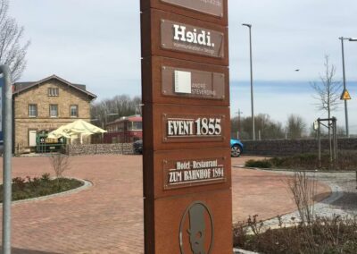 Großer brauner Wegweiser, der die Geschäfte in der Bahnhofstr. 61, einschließlich Restaurants und Agenturen, mit einem Gebäude, einem Parkplatz und einem bewölkten Himmel im Hintergrund.