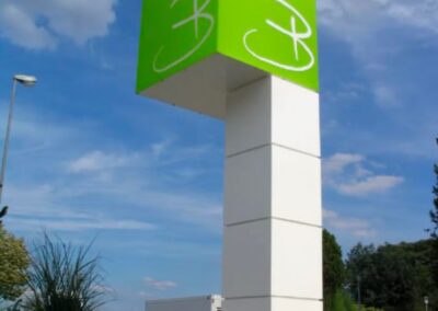 Ein hoher, weißer, rechteckiger Wegweiser mit einem grünen Würfel an der Spitze, der ein weißes abstraktes Logo zeigt, steht im Freien in der Nähe von Pflanzen und einer Straße.