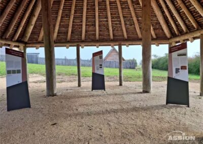 Drei Informationstafeln stehen unter einem großen, offenen Holzunterstand mit Strohdach; draußen sind eine Rasenfläche und ein Holzzaun zu sehen.