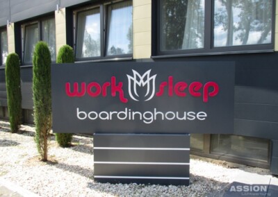 Außenschild mit der Aufschrift "Arbeitsschlaf-Pension" vor einem modernen Gebäude mit Fenstern, Sträuchern und Kiesflächen.