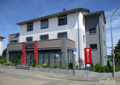 Modernes dreistöckiges Gebäude mit grauem und weißem Äußeren, großen Fenstern, einem roten Schild mit der Aufschrift "Novello" und umliegenden Grünflächen an einem sonnigen Tag.