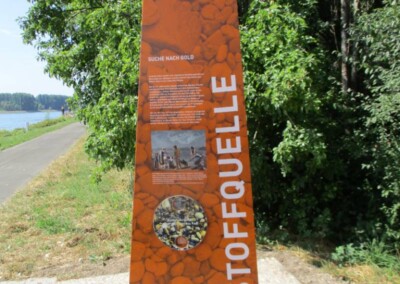 Ein großes orangefarbenes Informationsschild mit der Aufschrift "ROHSTOFFQUELLE" steht an einem gepflasterten Weg in der Nähe von Grünflächen und einem Fluss; das Schild enthält Text und Fotos über das Goldschürfen.