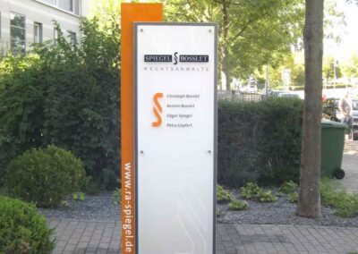 Auf einem Schild vor einem Büro ist die Anwaltskanzlei "Spiegel Bosselet Rechtsanwälte" mit vier Namen darunter und der Website der Kanzlei auf einer vertikalen orangefarbenen Tafel aufgeführt.