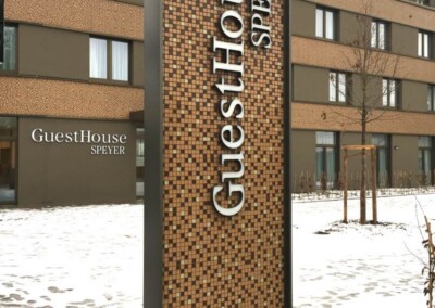 Großes Mosaikschild mit der Aufschrift "GuestHouse SPEYER" vor einem modernen Gebäude mit Schnee auf dem Boden. Das Gebäude zeigt den gleichen Text an seiner Wand.