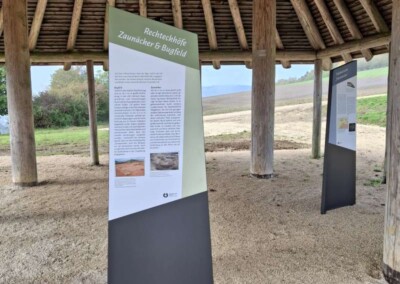 Zwei Informationstafeln stehen unter einem Holzpavillon mit Strohdach, der auf einer Kiesfläche steht, während im Hintergrund Felder und Bäume zu sehen sind.