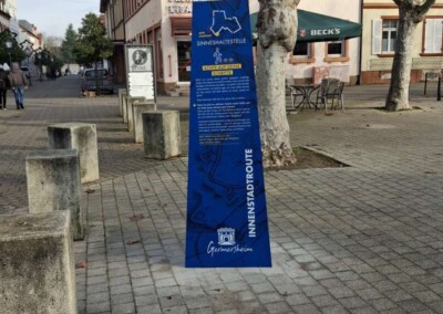 Ein blaues Hinweisschild mit der Aufschrift "Sinneshaltestelle" steht an einer gepflasterten Fußgängerstraße, die von Geschäften, Bäumen und Bänken gesäumt ist.