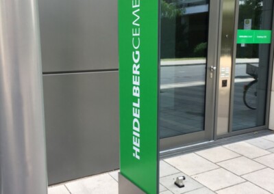Ein grünes vertikales Schild mit "HeidelbergCement" in weißen Buchstaben steht vor dem Eingang eines modernen Bürogebäudes.