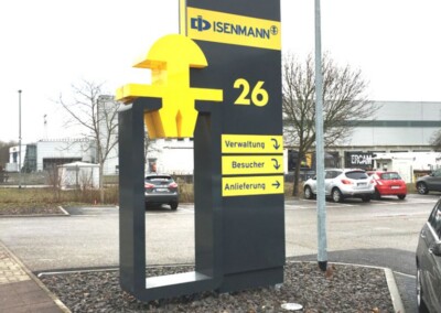 Großes Außenschild für Isenmann an der Adresse 26, mit fettem gelben Logo und Pfeilen, die zur Verwaltung, zum Besucher und zur Anlieferung führen. Das Schild steht auf einer Kiesfläche in der Nähe geparkter Autos.