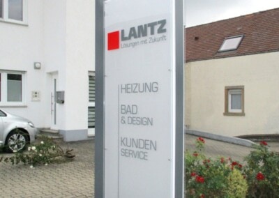 Auf einem hohen Wegweiser vor einem Gebäude steht "Weinstraße 62", das LANTZ-Firmenlogo und Dienstleistungen: Heizung, Bad & Design, Kundendienst. In der Nähe sind Sträucher und Pflastersteine zu sehen.