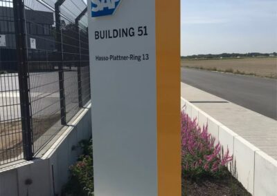 Ein Schild weist auf das SAP-Gebäude 51 am Hasso-Plattner-Ring 13 hin. Es steht in der Nähe eines Zauns und einer Straße mit einigen Blumen in der Nähe.