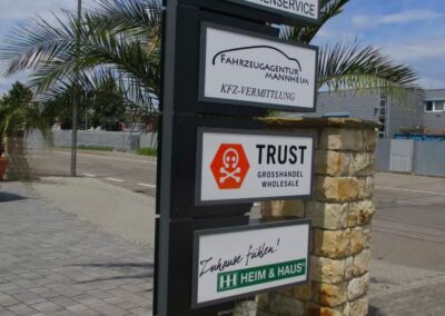 Auf einem hohen Wegweiser stehen neben einer Palme und einer Steinsäule an einer gepflasterten Straße die Namen von Unternehmen wie Limousinenservice, Kfz-Agentur, Treuhandgroßhandel und Heimservice.