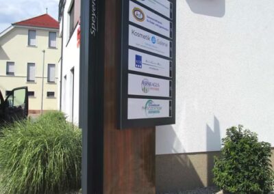 Vor einem Gebäude mit einem großen roten Logo an der Wand steht ein Schild mit einer Auflistung von Unternehmen; in der Nähe gibt es Pflanzen und Steine in der Landschaft.