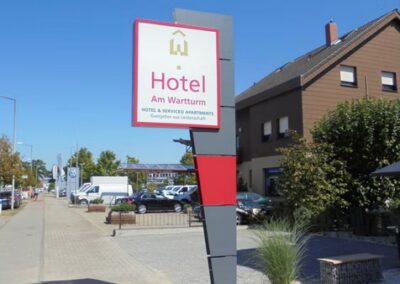 Ein Bürgersteig mit einem modernen Schild für "Hotel Am Warturm" neben einer Straße und einem braunen Gebäude, mit geparkten Autos im Hintergrund.