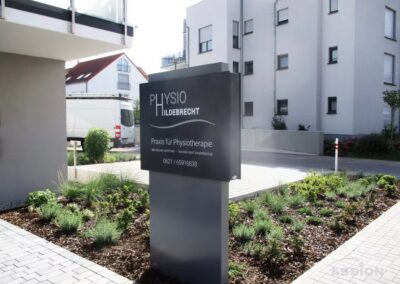 Ein modernes Schild für die Physiotherapieklinik Physio Hildebrecht steht vor einem Gebäude, mit anderen Wohngebäuden und einem geparkten Lieferwagen im Hintergrund.