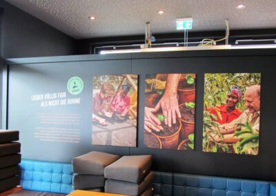Drei Fotos an einer Wand zeigen Menschen beim Pflanzen und Pflegen von Kaffeepflanzen, mit deutschem Text und gestapelten Sitzkissen im Vordergrund.