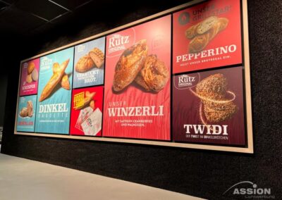 An einer Wand hängen fünf bunte Plakate, die für verschiedene Brotsorten und Backwaren der Marke "Rutz Original" werben, mit Beschreibungen in deutscher Sprache.