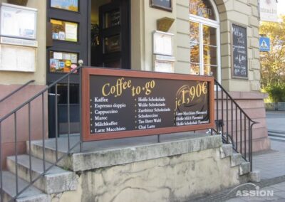 Der Eingang eines Cafés mit einem großen Schild, auf dem verschiedene Kaffee- und Heißgetränke zum Mitnehmen zum Preis von je 1,90 € in deutscher und englischer Sprache aufgelistet sind.