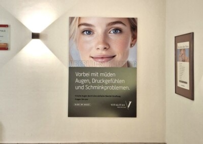 In einem Flur mit gefliestem Boden ist ein großes Plakat mit dem Gesicht einer Frau und einem deutschen Text über Ermüdungserscheinungen der Augen zu sehen; darüber befindet sich ein Notausgangsschild.