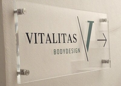 Ein durchsichtiges Acrylschild an einer Wand zeigt "Vitalitas Bodydesign" mit einem großen "V" und einem nach rechts zeigenden Pfeil. In der Ecke steht das Logo "Assion Lichtwerbung".