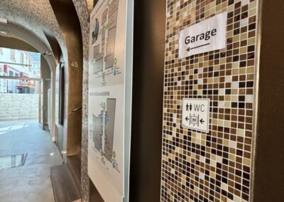 Innenflur mit braunen Mosaikfliesen, einem an der Wand befestigten Lageplan und Schildern, die den Weg zur Garage und zu den Toiletten weisen.