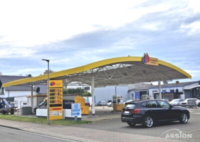 Eine kleine Tankstelle mit einem gelben Vordach, einem Benzinpreisschild und einem schwarzen Auto, das in der Nähe geparkt ist. Die Tankstelle liegt an einer ruhigen Straße mit Gebäuden und Bäumen im Hintergrund.