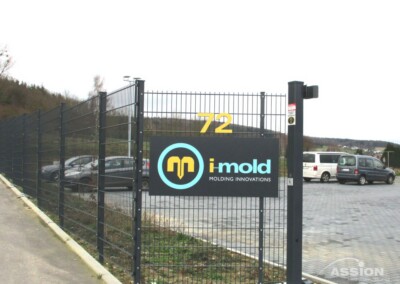 Ein schwarzer Metallzaun mit einem Tor zeigt ein Schild mit der Aufschrift "imold Molding Innovations" und der Nummer 72; auf dem Parkplatz hinter dem Zaun sind parkende Autos zu sehen.