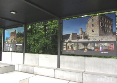 Auf Schautafeln im Freien werden Fotos und Informationen über ein historisches Steingebäude gezeigt, das von grünen Bäumen und Sitzgelegenheiten aus Beton eingerahmt wird.