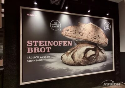 Werbeplakat für "Steinofen Brot" mit zwei Broten und dem Text, dass es aus Dinkel und Roggen hergestellt wird und täglich außer sonntags erhältlich ist.