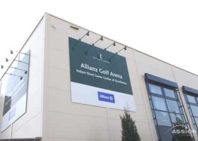 Auf einem großen Schild an einem Gebäude steht: "Allianz Golf Arena Indoor Short Game Center of Excellence" mit Allianz-Logos und sichtbaren Außenfenstern.