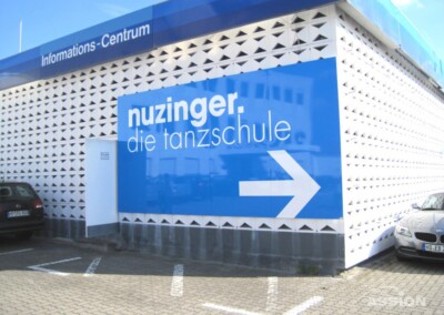 Auf einem großen blauen Schild an einem weißen Gebäude steht "nuzinger. die tanzschule" mit einem weißen Pfeil, der nach rechts zeigt. Über dem Gebäude steht "Informations-Centrum". In der Nähe sind Autos geparkt.