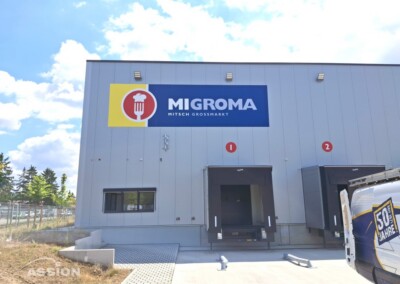 Außenansicht des Lagers mit dem "Migroma"-Schild über zwei Laderampen mit den Nummern 1 und 2; rechts ist teilweise ein Lieferwagen mit dem Logo "50 Jahre" zu sehen.