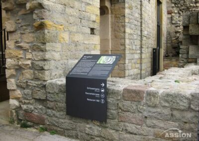 Informationsschild auf einer Steinmauer an einer historischen Stätte, mit Text und Symbolen, die den Weg zu Eintrittskarten, Toiletten und Restaurant anzeigen.