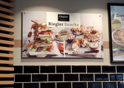 Eine Wandkarte zeigt Bilder von Sandwiches, Bagels und Dips unter der Überschrift "Riegler Snacks" mit einem Slogan in deutscher Sprache über einer schwarz-weiß gekachelten Wand.