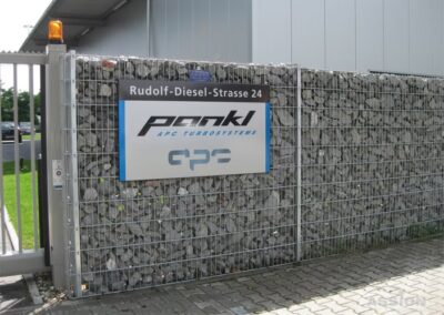 Ein Metallzaun mit einem Schild mit der Aufschrift "Rudolf-Diesel-Straße 24" und den Logos von Penk APC Turbosystems und APC neben einem Gebäude.