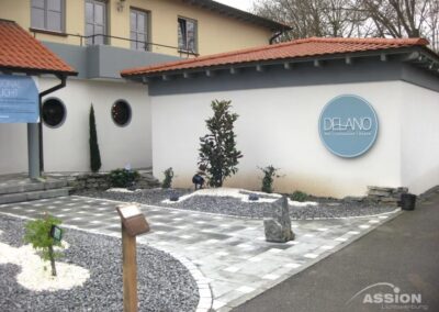 Modernes Gebäude mit einem runden "Delano"-Schild an einer weißen Wand, begrünt mit Kies, kleinen Pflanzen und einem gepflasterten Gehweg.