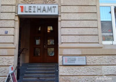 Steinerner Gebäudeeingang mit einem Schild über der Tür mit der Aufschrift "Leihamt" und einem weiteren Schild rechts neben der Tür mit der Aufschrift "Städtisches Leihamt Mannheim". Eine Treppe führt zu einer hölzernen Doppeltür.