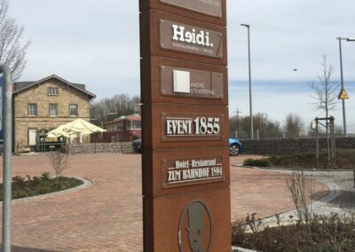 Ein hoher brauner Wegweiser listet mehrere Geschäfte, darunter Restaurants und Büros, vor einem Steingebäude und einem Parkplatz in der Bahnhofstr. 61.