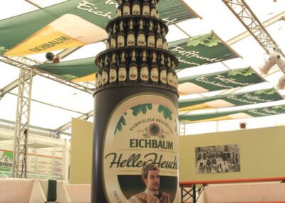 In einem Bierzelt mit Tischen und Bänken steht eine große flaschenförmige Auslage mit der Aufschrift "Eichbaum Helles Leucht", die mit Reihen kleinerer Flaschen geschmückt ist.