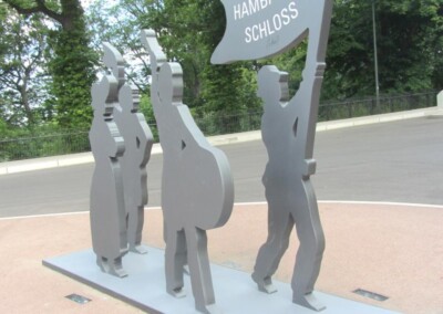 Eine graue Metallskulptur mit vier Personen, von denen eine eine Fahne mit der Aufschrift "Hambacher Schloss" hält, steht auf einer gepflasterten Fläche mit Bäumen und einer Straße im Hintergrund.
